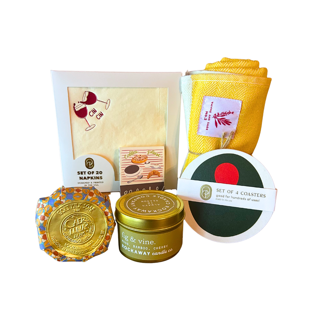 Collection of home decor items including candle, coasters, napkins, dish towel, matchbook, and goat milk soap.