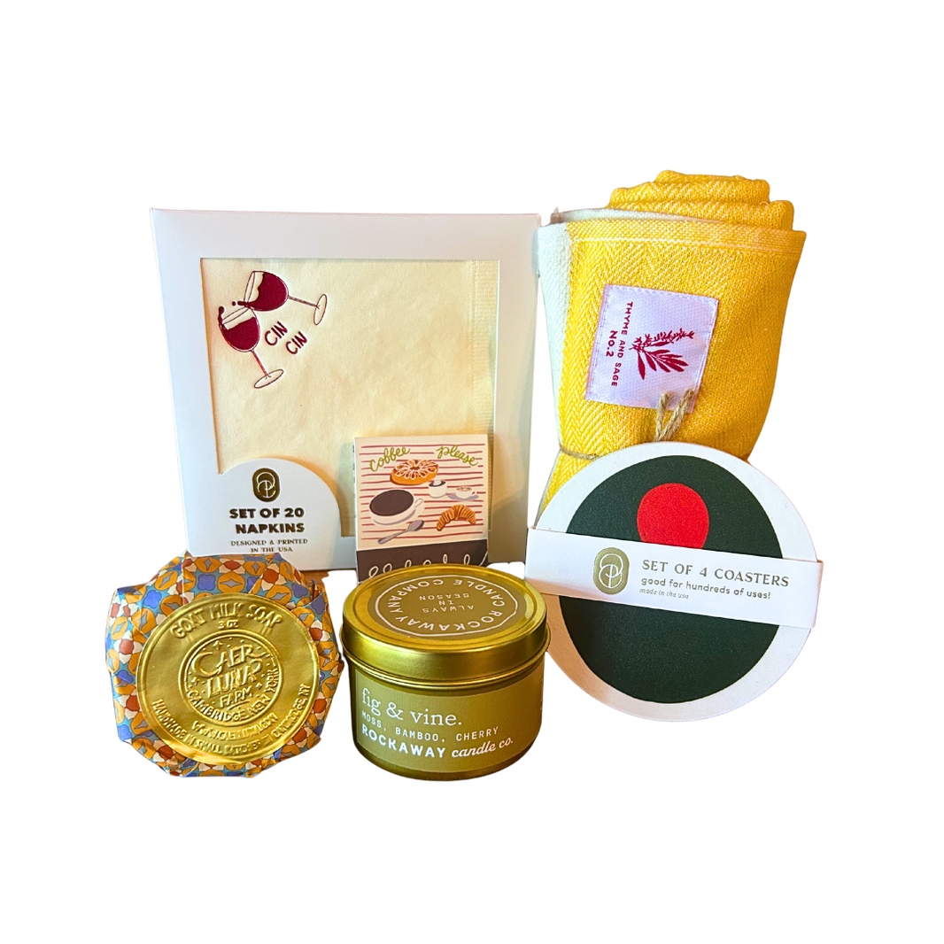 Collection of home decor items including candle, coasters, napkins, dish towel, matchbook, and goat milk soap.